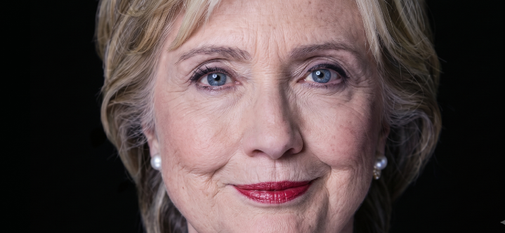 A close-up portrait of Hillary Clinton with a gentle smile, wearing pearl earrings and a dark top, set against a solid black background.