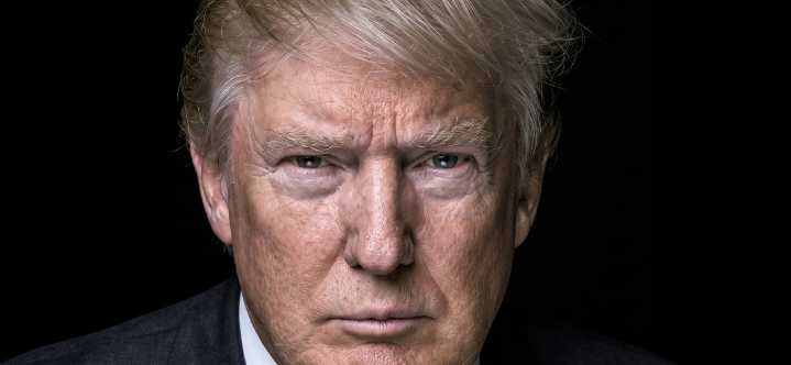 A close-up, dramatic portrait of Donald Trump looking directly at the camera with a serious expression against a solid black background.